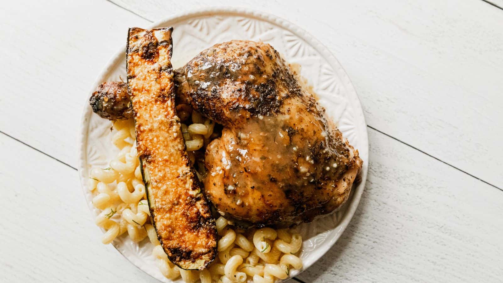 A plate with baked chicken, a piece of roasted zucchini, and cooked curly pasta on a white wooden table.