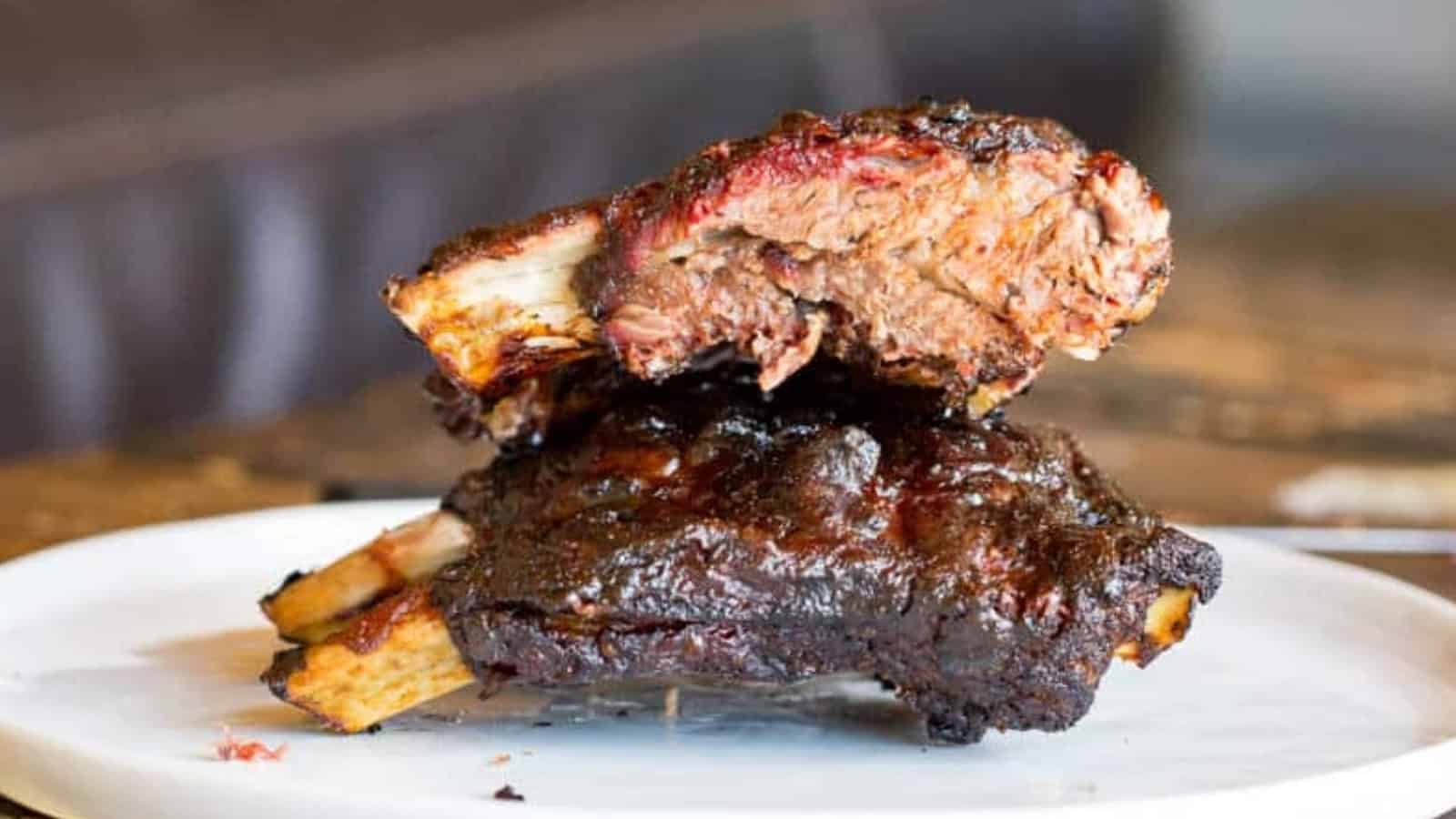Two stacked, saucy smoked beef ribs on a white plate, showing a juicy and tender interior.