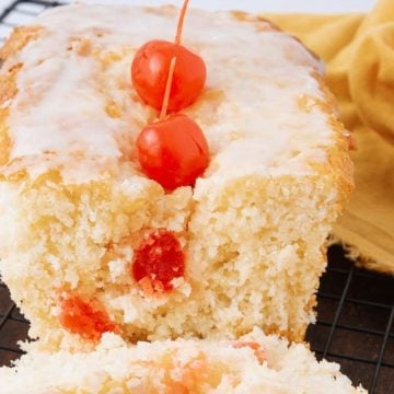 A sliced loaf cake with icing and cherries on top, sitting on a cooling rack beside a yellow cloth.