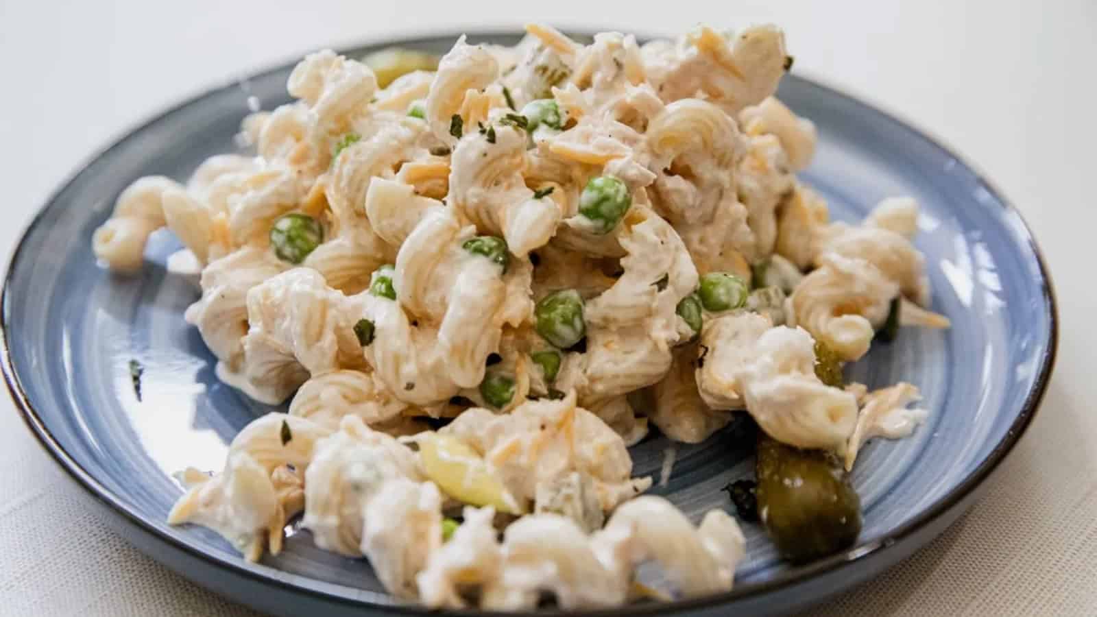 A plate of creamy pasta salad with green peas, herbs, and a small pickle on a blue dish.