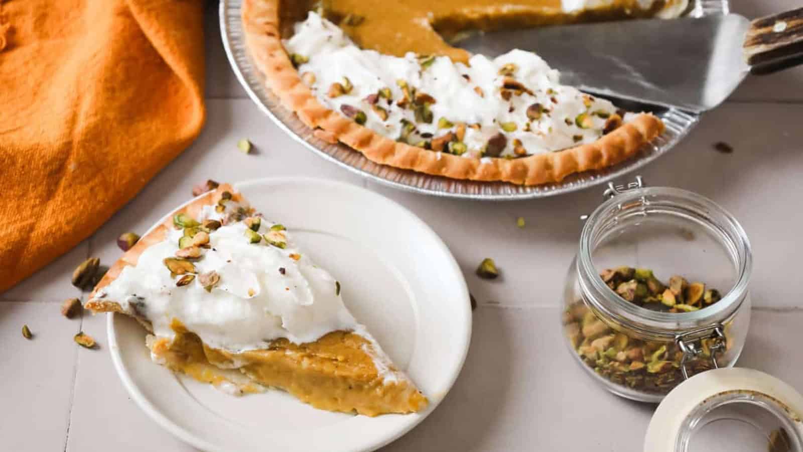 A slice of pumpkin pie with whipped cream and chopped pistachios on a plate, next to the rest of the pie.