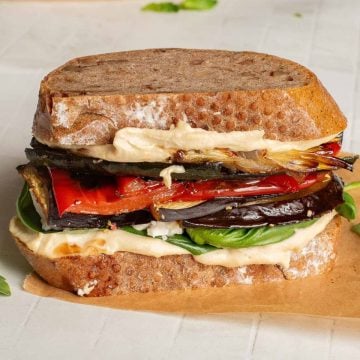 A sandwich with roasted vegetables, leafy greens, and hummus on rustic bread, placed on parchment paper.