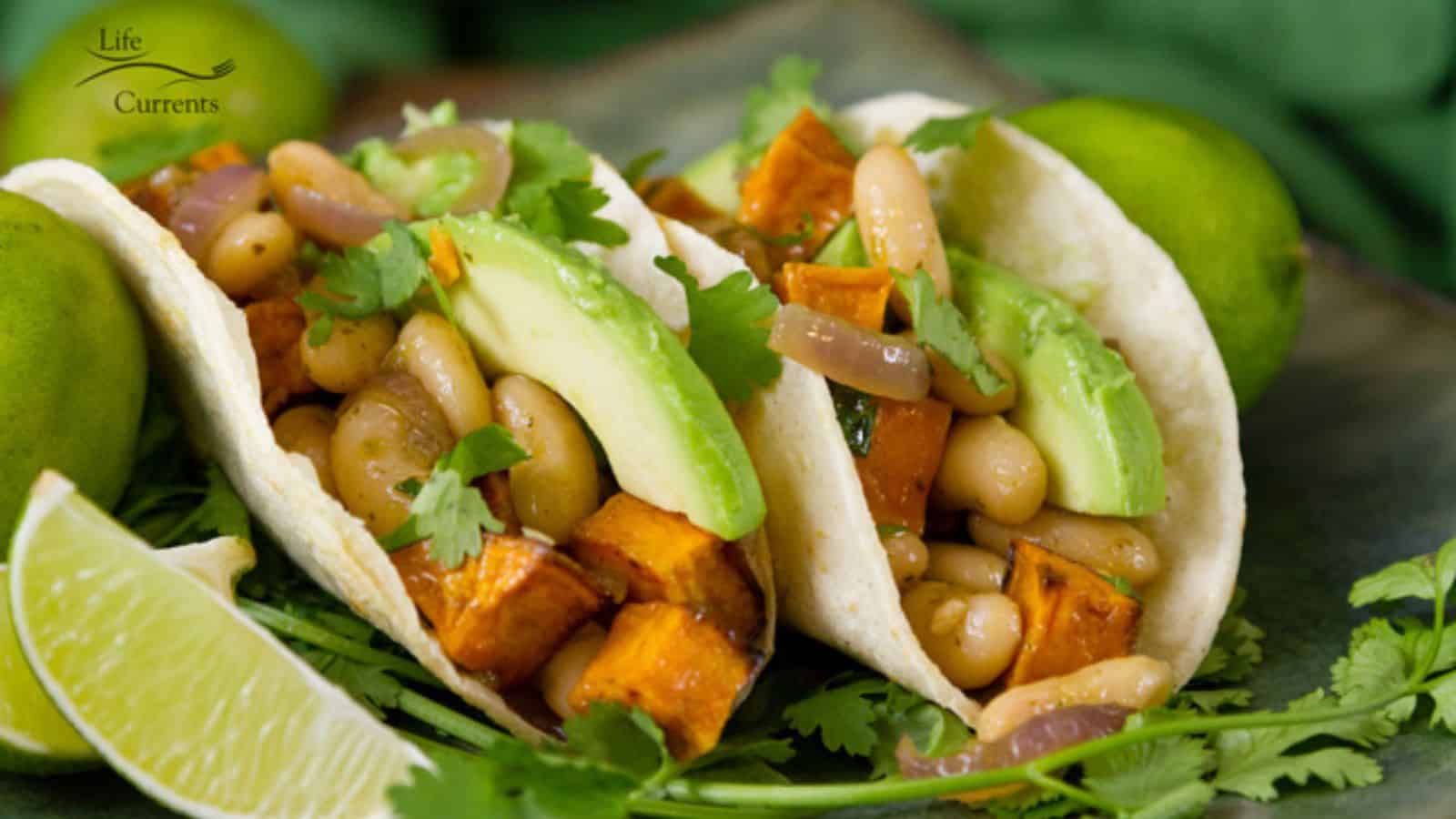 Two veggie tacos with avocado, beans, sweet potatoes, onions, and cilantro, garnished with lime wedges.