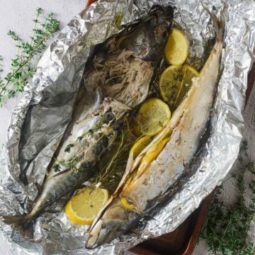 Two baked whole fish on foil with lemon slices and fresh herbs, partially flaked open to reveal the inside.