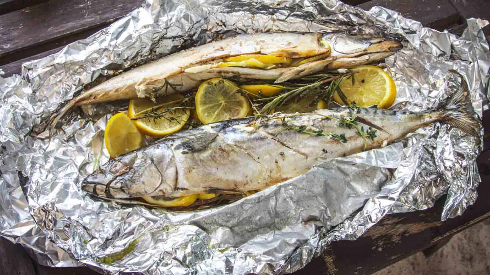 Two whole fish baked in foil with lemon slices and herbs placed on a rustic wooden surface.