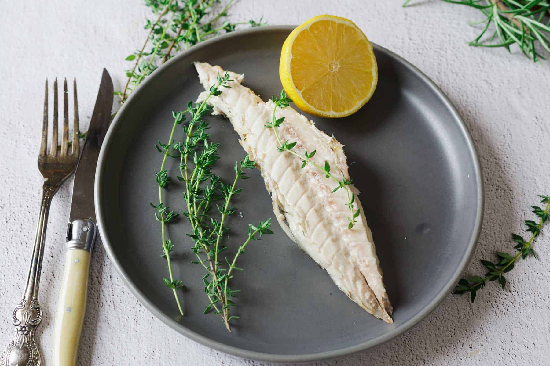 A cooked fish fillet with fresh thyme and a lemon half on a gray plate, with a fork and knife beside it.