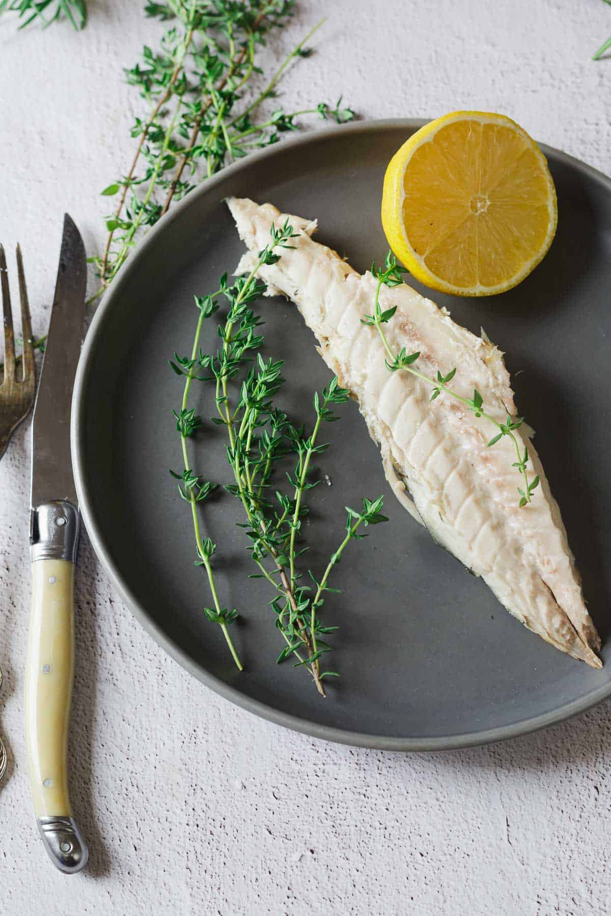 Cooked fish fillet with fresh thyme and half a lemon on a dark plate, next to a fork and knife.