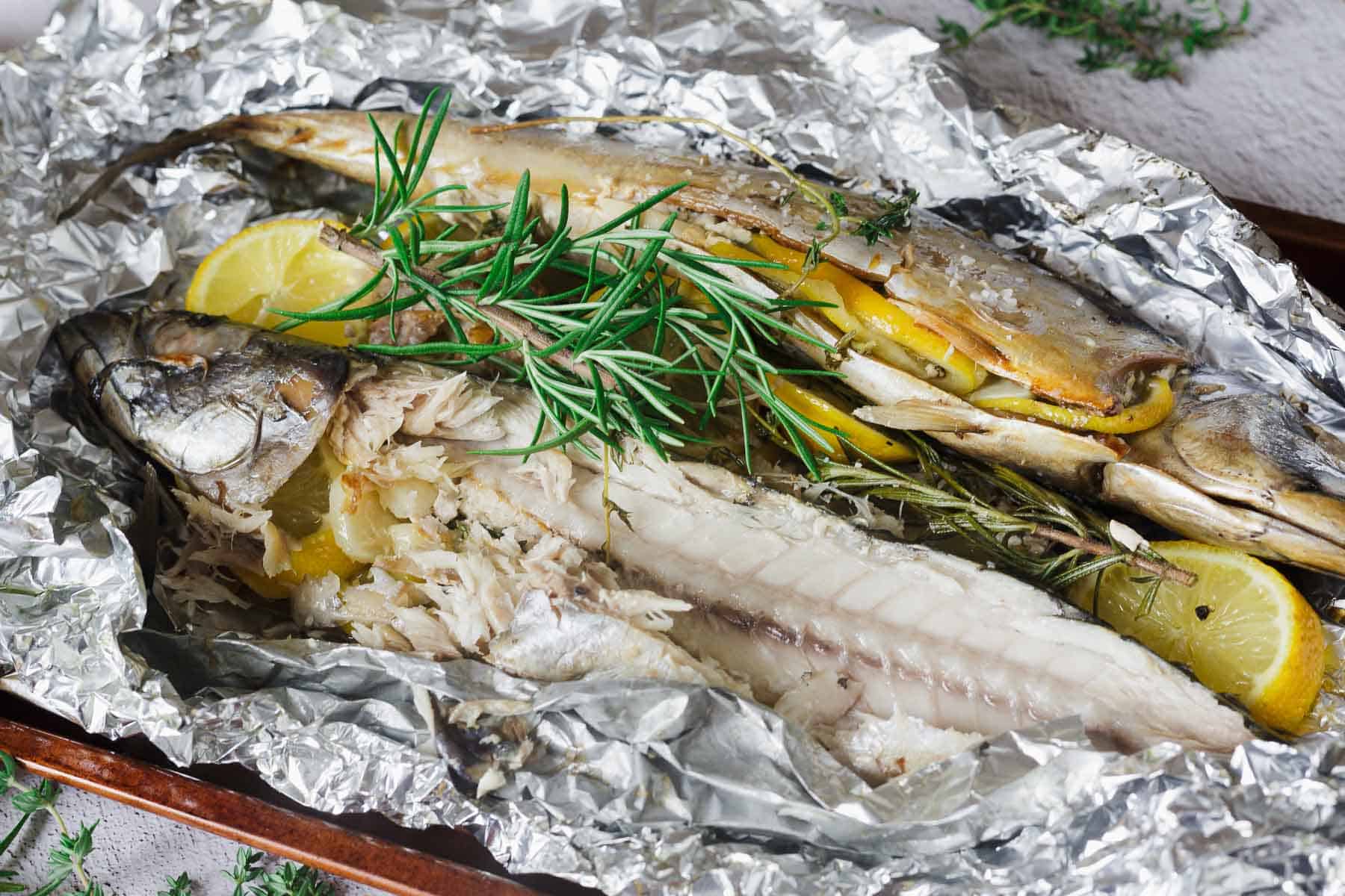 Two cooked whole fish with lemon slices and rosemary, served on aluminum foil.