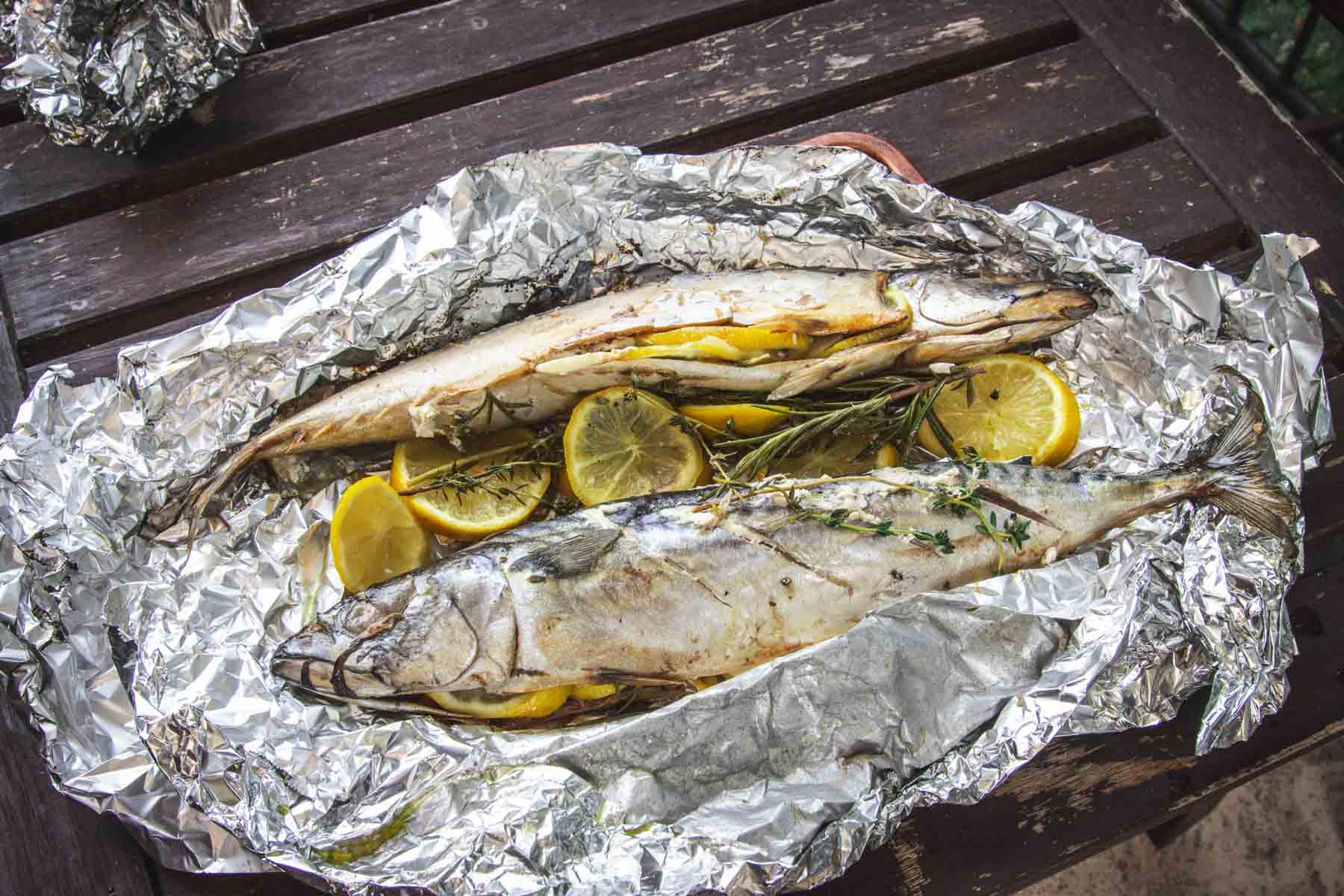 Two whole cooked fish with lemon slices and herbs on aluminum foil, on a wooden table.