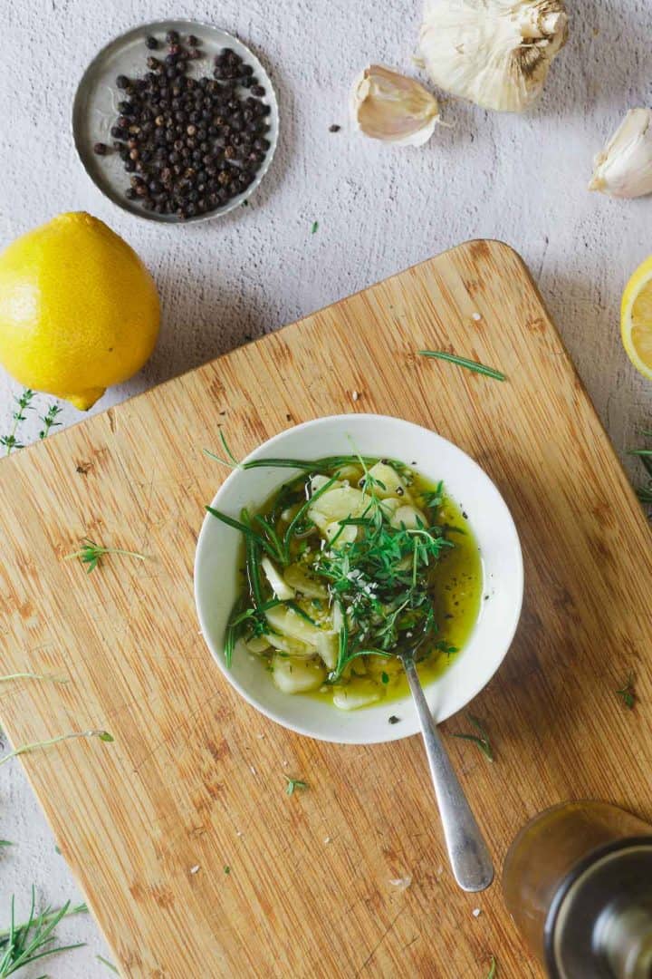 Bowl of herb-infused olive oil on a cutting board with lemon, garlic, and black pepper nearby.