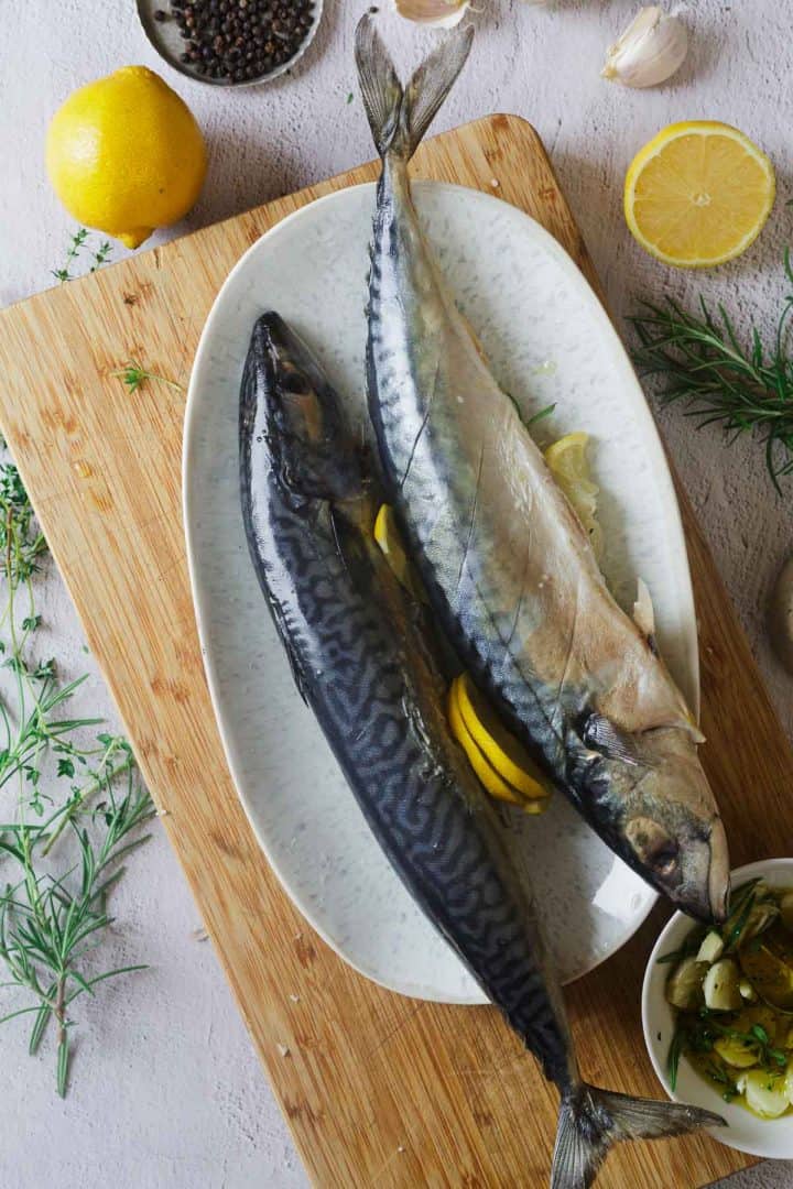 Two raw mackerel fish on a white plate with lemon slices, herbs, and spices on a wooden board.