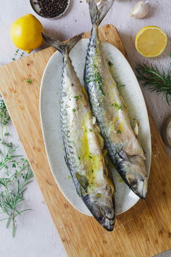 Two whole raw fish with herbs and lemon on a white oval plate, placed on a wooden cutting board.