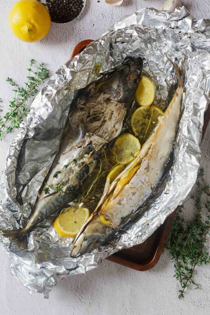 Two whole baked fish with lemon slices and herbs in aluminum foil on a white surface.