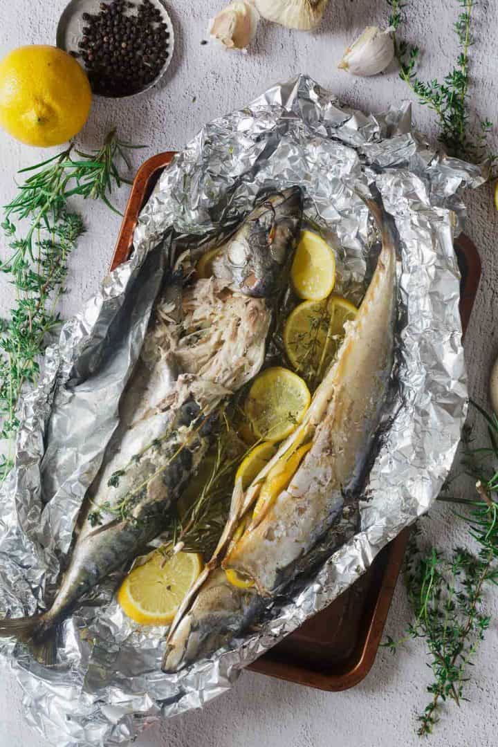 Two baked whole fish with lemon slices and herbs in foil on a tray, surrounded by fresh herbs, garlic, and pepper.