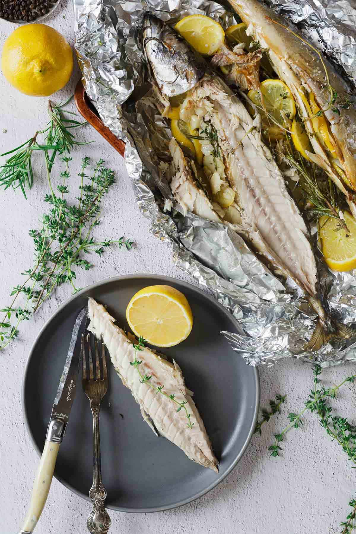 Baked fish fillet on a plate with lemon; whole baked fish with herbs and lemon in foil nearby.