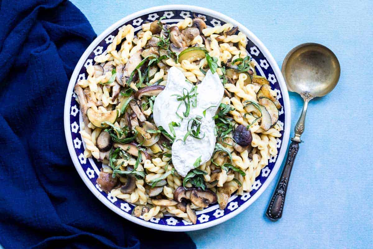 Pasta with mushrooms, zucchini, burrata, and fresh basil served in a patterned bowl, next to a gold spoon.