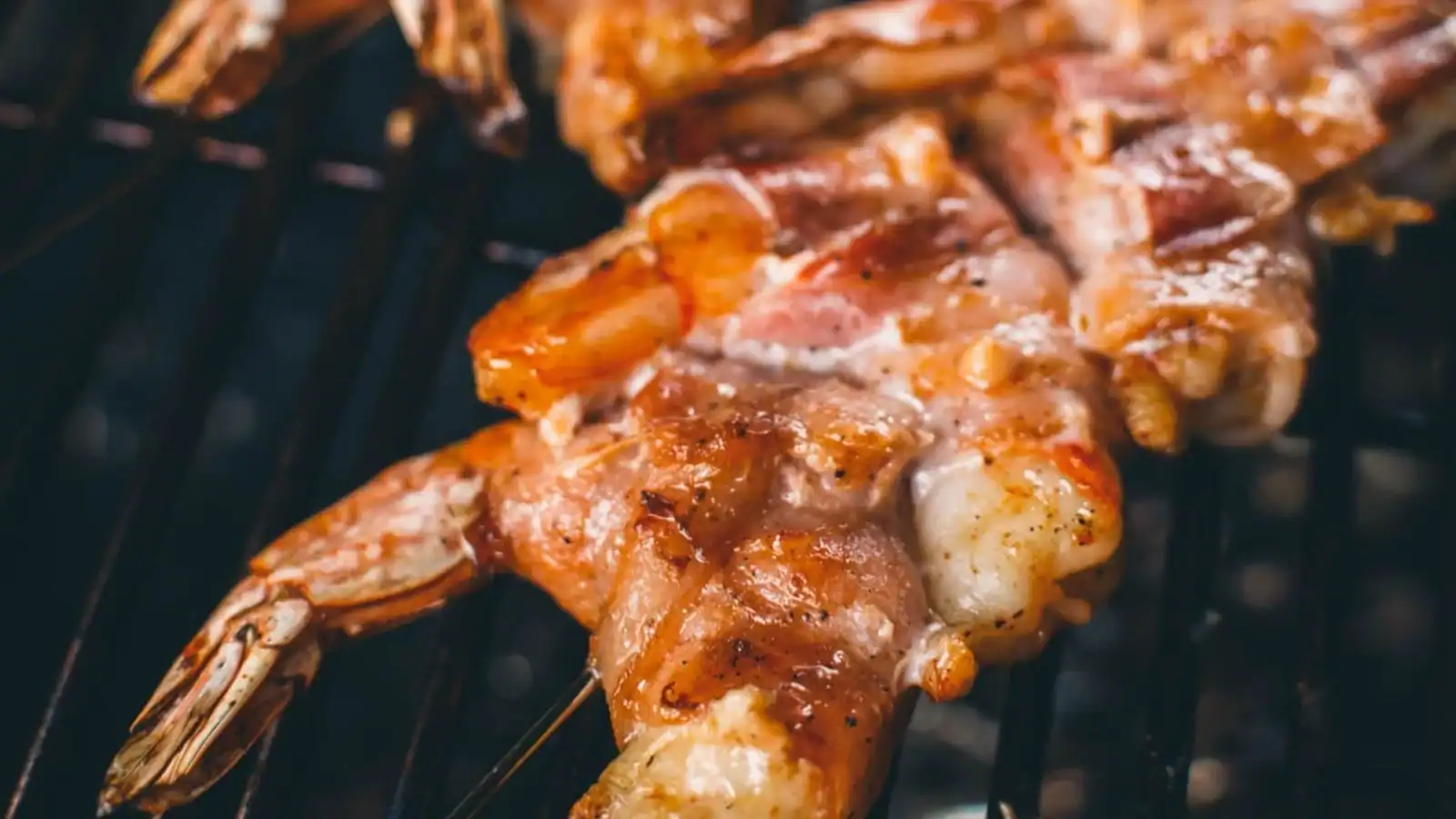 Grilled bacon-wrapped shrimp with crisped bacon exterior plated next to dipping sauce.