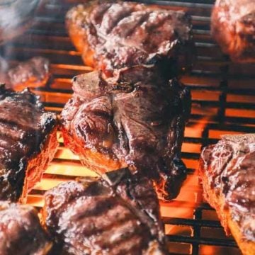 Several pieces of steak are grilling over an open flame, producing smoke and seared grill marks.
