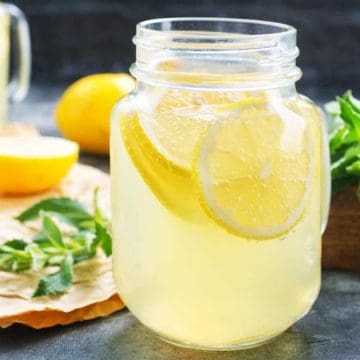 A mason jar of lemonade with lemon slices, fresh lemons, and mint leaves on a dark table.