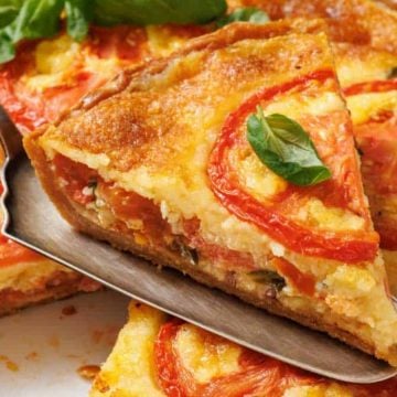 A slice of tomato and cheese quiche with basil being lifted from the whole quiche on a metal spatula.