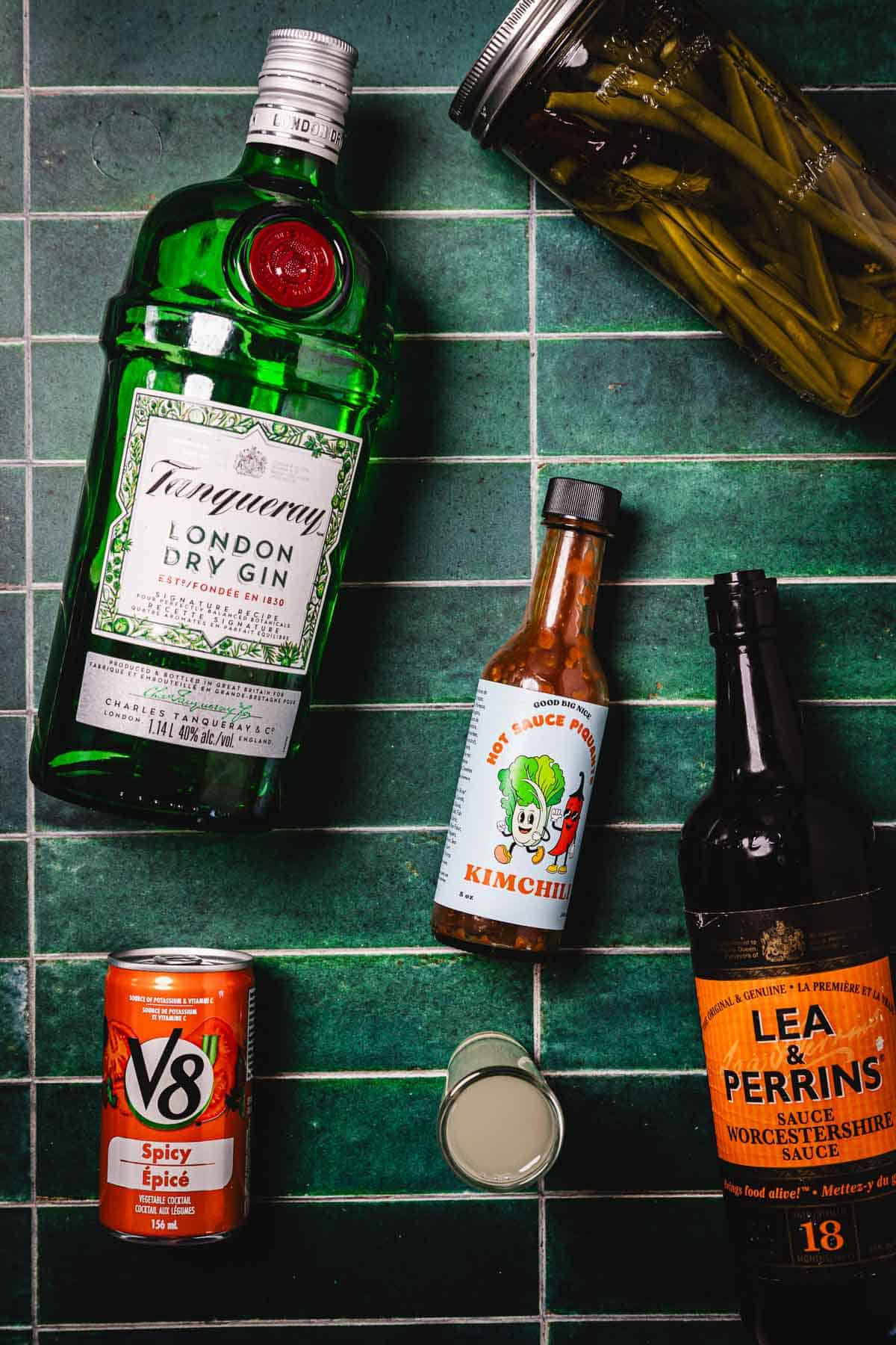 Tanqueray gin, V8 can, kimchi hot sauce, Worcestershire sauce, green beans jar, and shot glass on green tiles—everything you need for a bold Caesar Cocktail awaits.