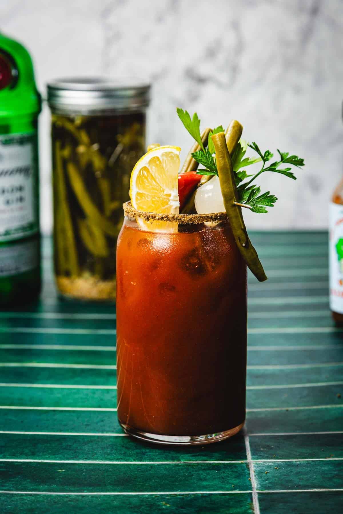 A Bloody Mary cocktail, reminiscent of a classic Caesar Cocktail, garnished with lemon, olive, green beans, celery, and parsley on a green tile surface.