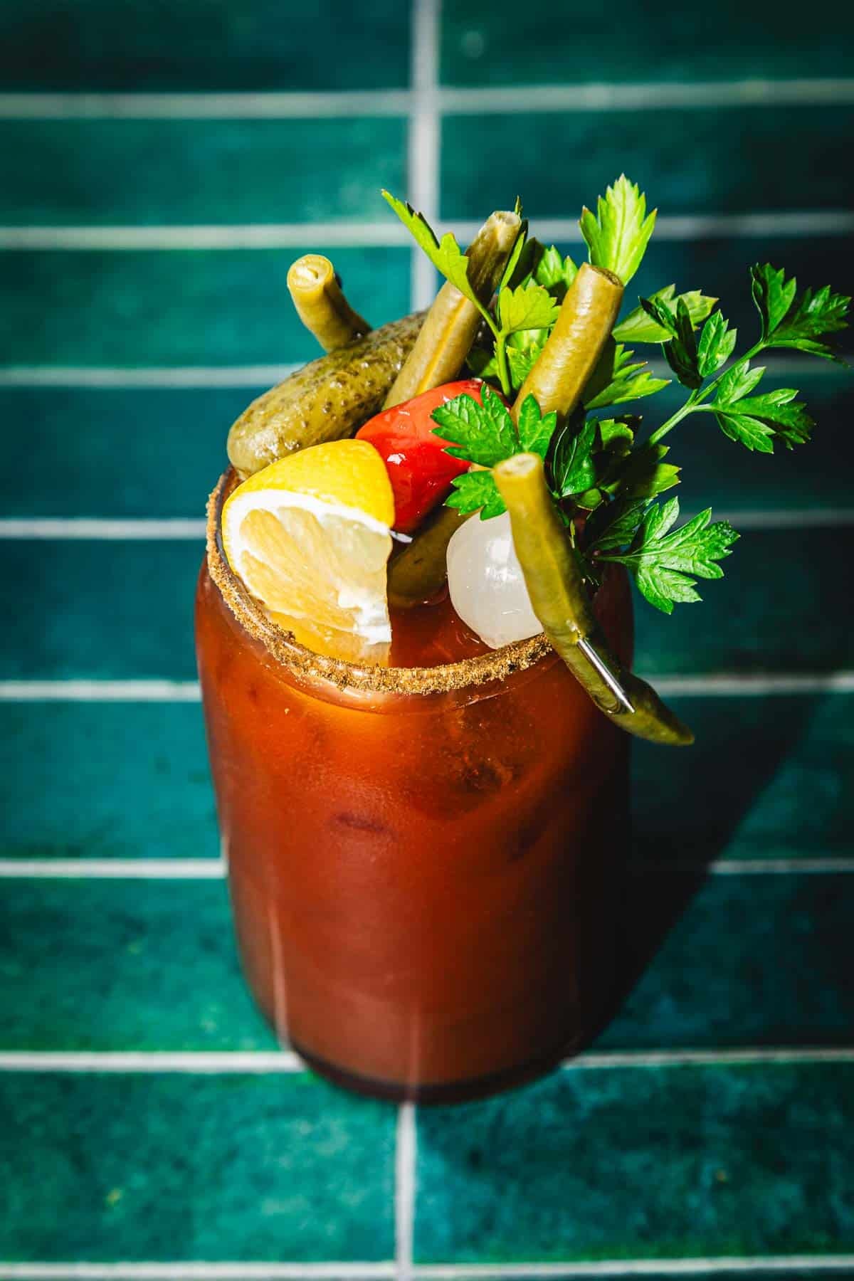 A Bloody Mary cocktail, reminiscent of a classic Caesar Cocktail, garnished with lemon, pickles, green beans, olives, and parsley on a teal tile surface.
