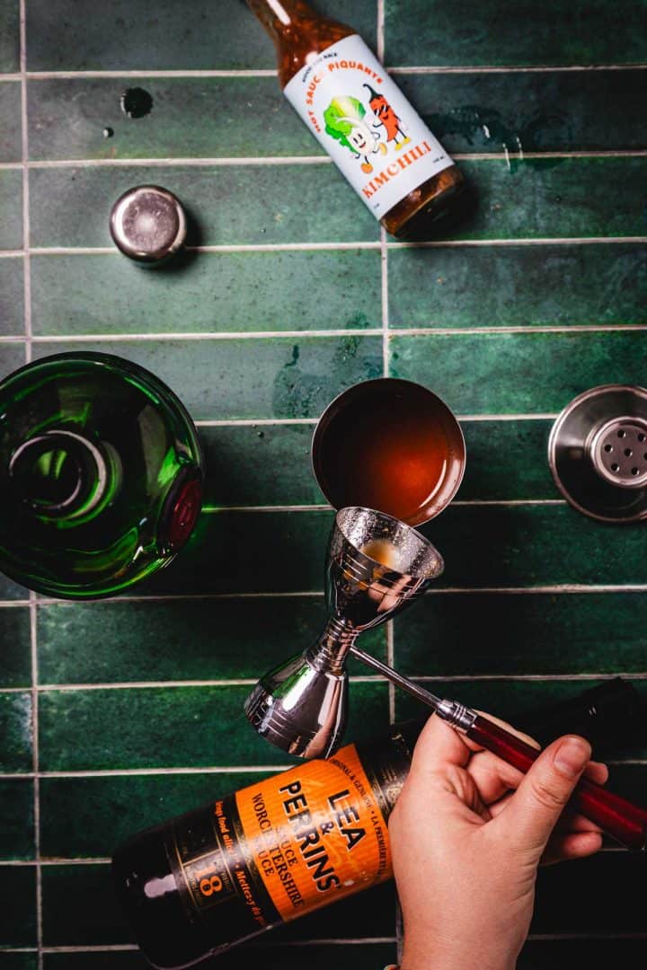 Hand measuring sauce into jigger for a Caesar Cocktail, with hot sauce, shaker, and green bottle on a green tiled surface.