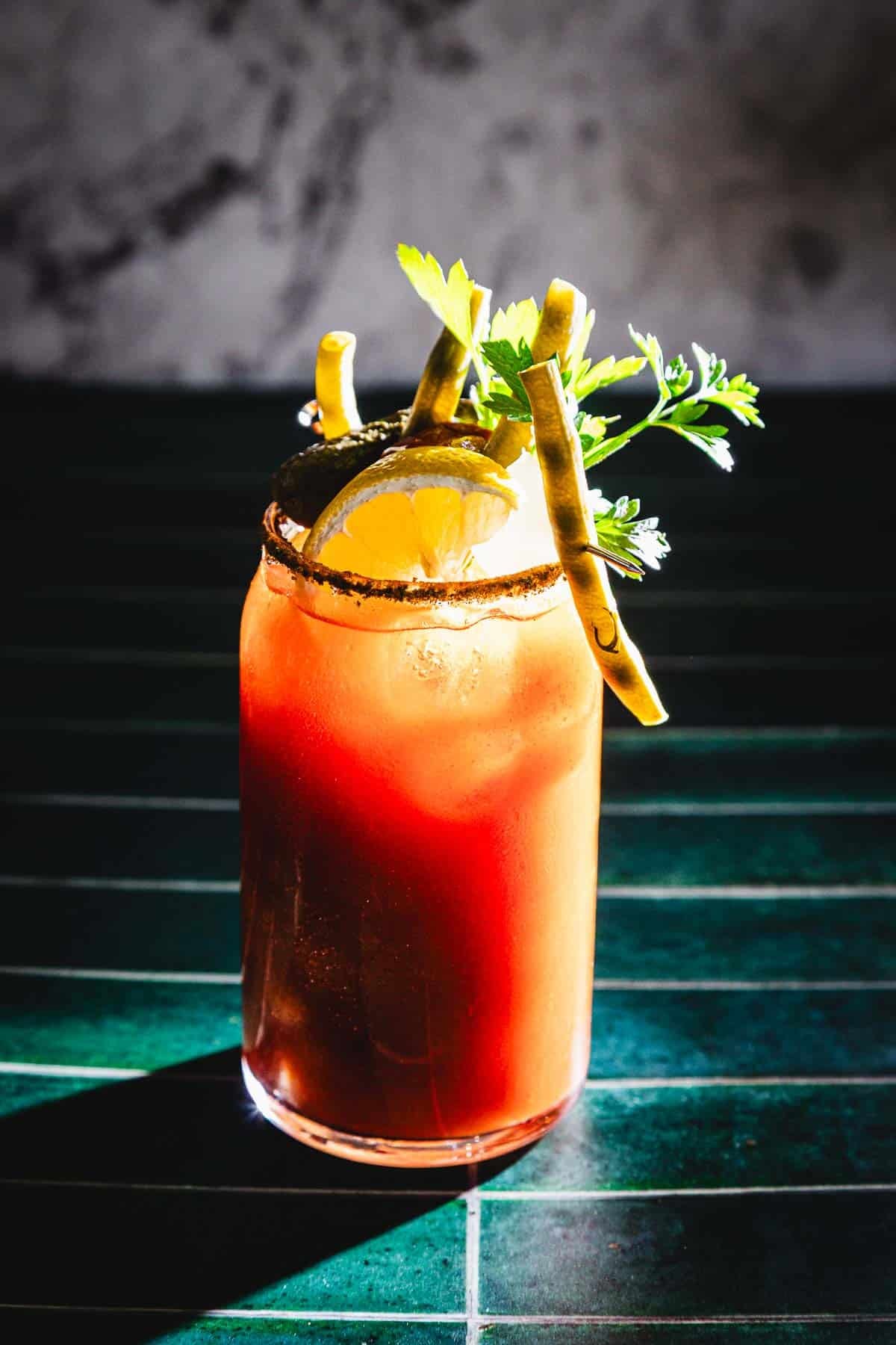 A Bloody Mary cocktail, reminiscent of a classic Caesar Cocktail, garnished with lemon, celery, green herbs, and two breadsticks on a green tiled surface.