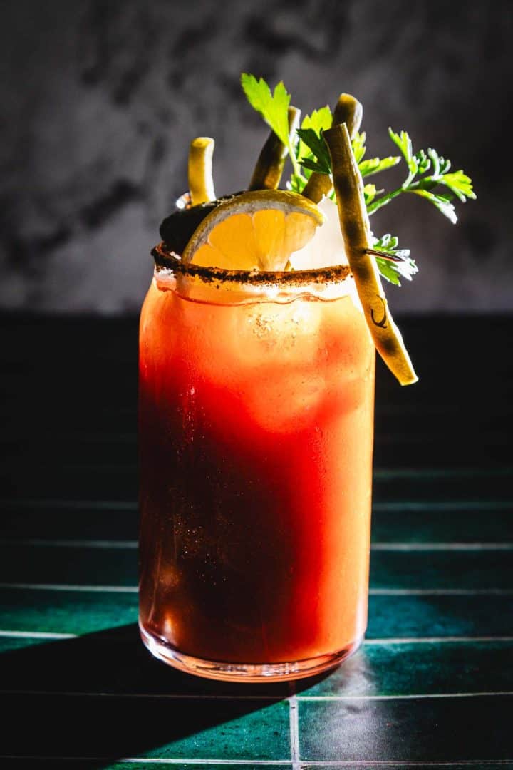 A Caesar Cocktail with ice, lemon slice, green beans, and herbs is served in a tall glass on a green surface.