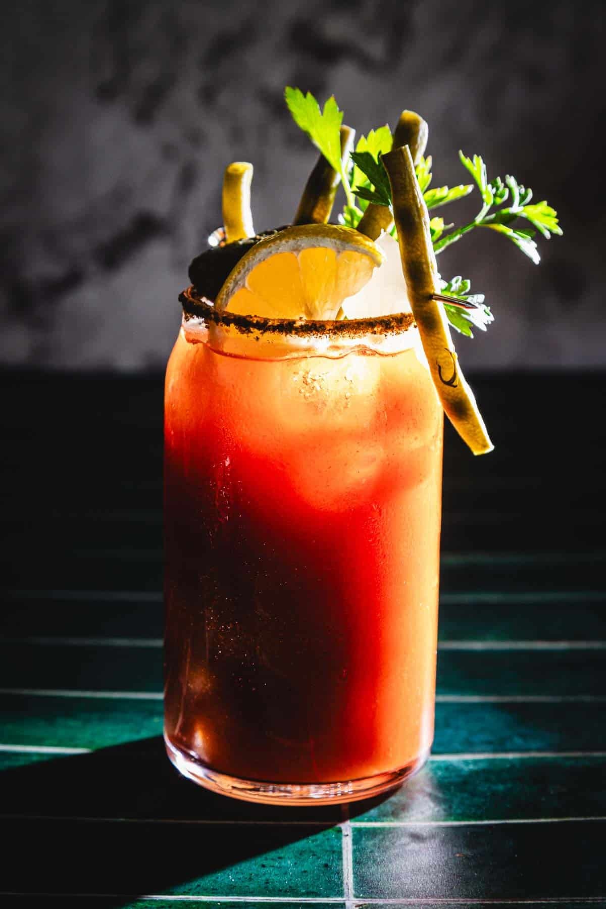 A Caesar Cocktail with ice, lemon slice, green beans, and herbs is served in a tall glass on a green surface.