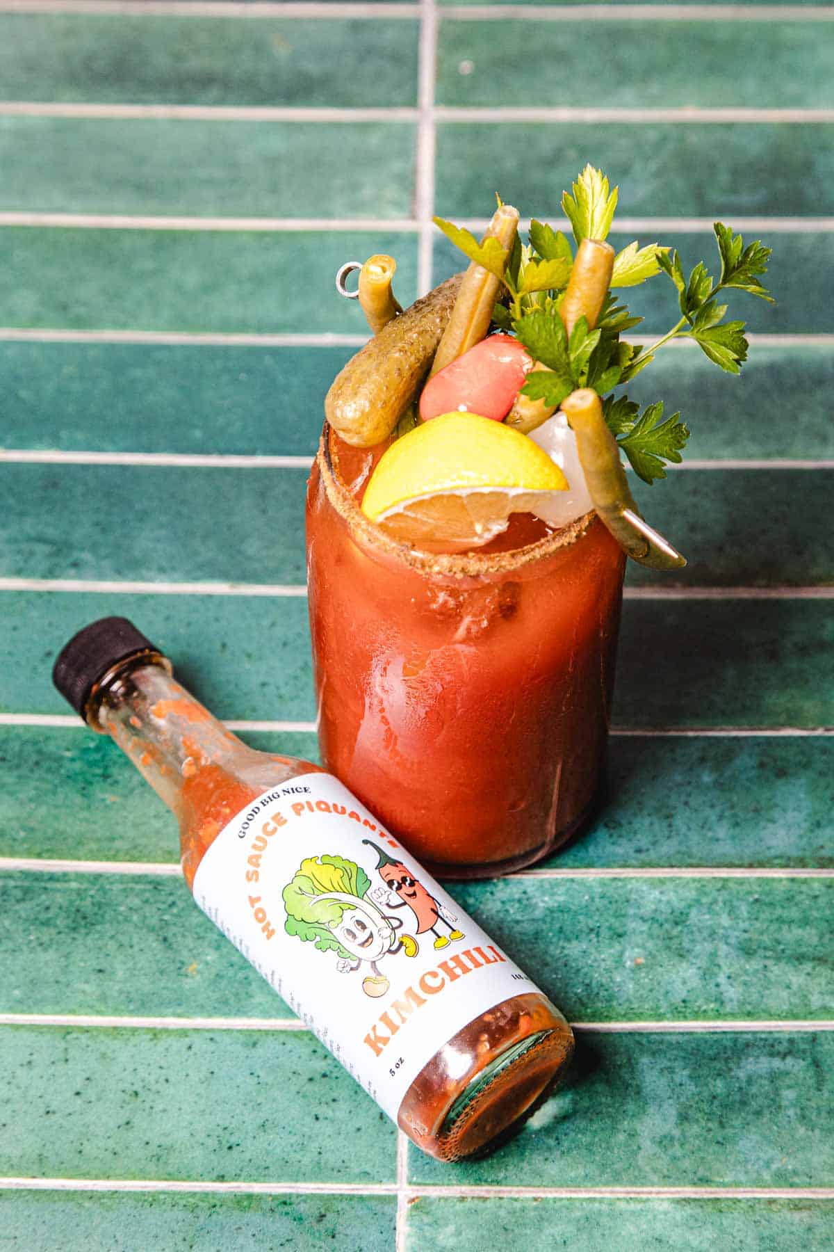 A Caesar Cocktail garnished with veggies and lemon sits next to a bottle of hot sauce on a green tiled surface.