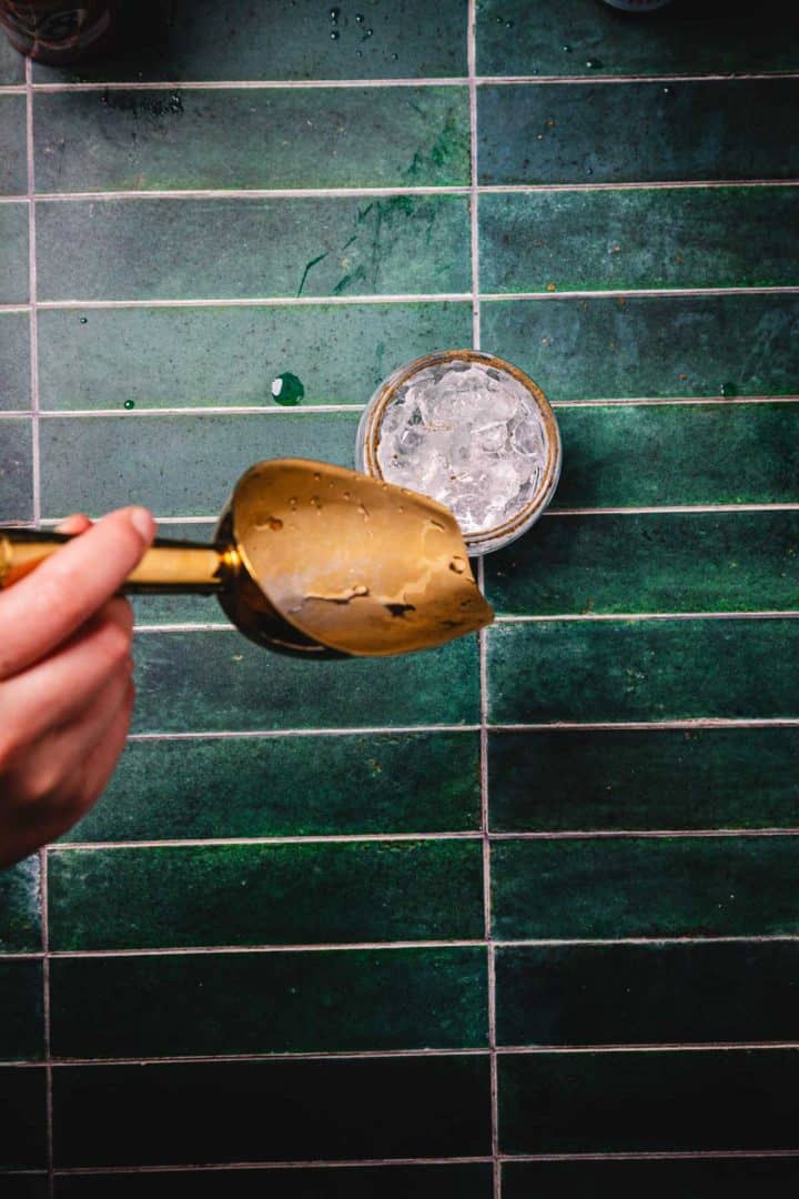 Hand holding a large brass ice scoop above a glass filled with ice on green tiled surface, ready to craft the perfect Caesar Cocktail.