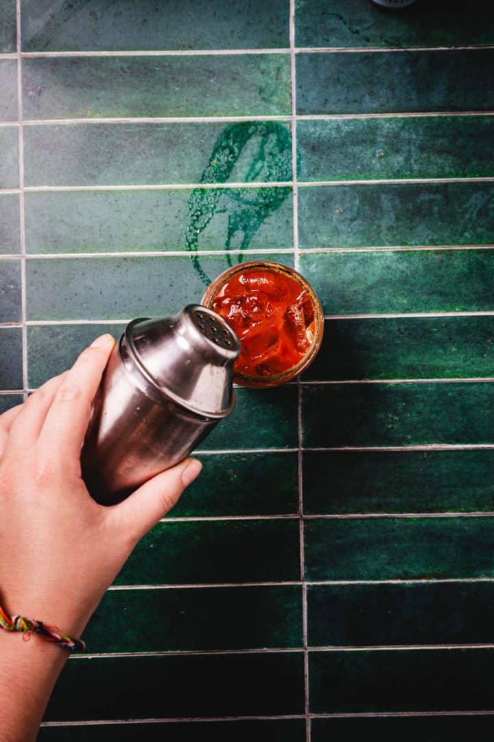 Hand holding a cocktail shaker above a glass with a red Caesar Cocktail and ice on a green tiled surface.