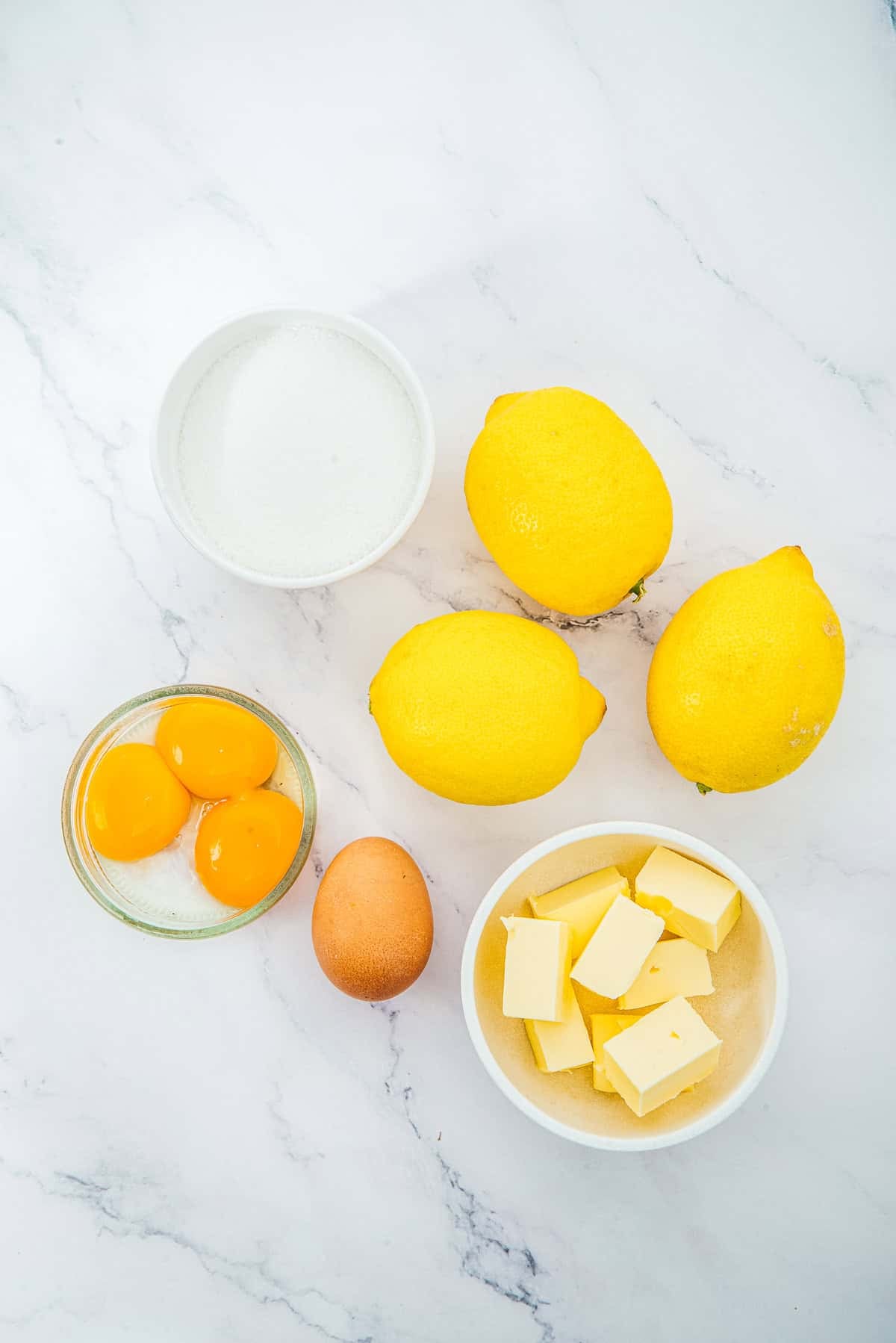 Three lemons, butter cubes, sugar, egg yolks, and a whole egg rest on a white marble surface—ready to be transformed into the luscious filling for delightful Lemon Cheesecake Trifle Cups.