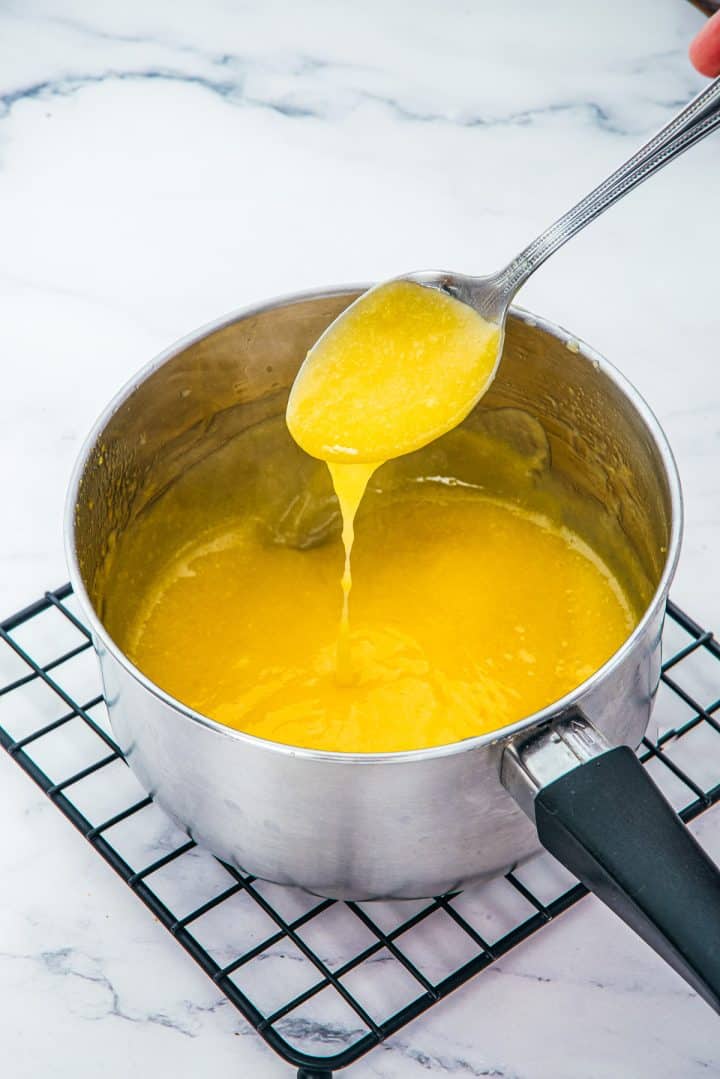 A spoon holds yellow sauce-perfect for Lemon Cheesecake Trifle Cups-dripping into a saucepan on a cooling rack, with a marble background.