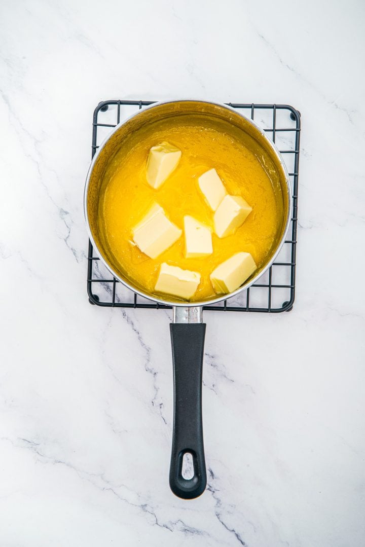 A saucepan with melted butter and butter cubes, on a cooling rack over a white marble surface-perfect for preparing the delicious base of Lemon Cheesecake Trifle Cups.