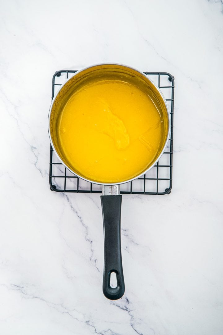 A saucepan with yellow sauce, perfect for making Lemon Cheesecake Trifle Cups, sits on a cooling rack on a white marble surface.