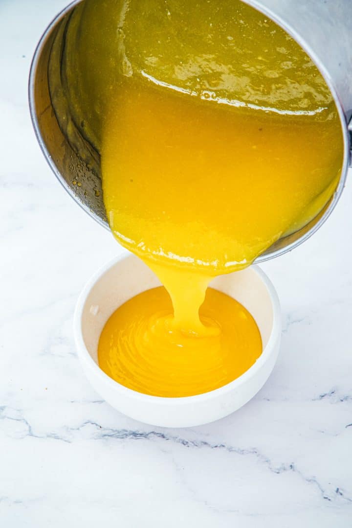 Bright yellow liquid, reminiscent of the luscious filling in Lemon Cheesecake Trifle Cups, being poured from a metal pot into a white bowl on a marble surface.