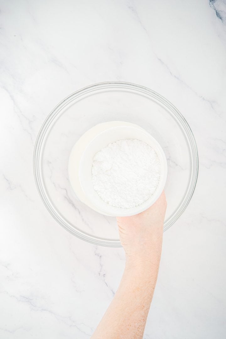 A hand holds a white bowl of powdered sugar above an empty glass mixing bowl on a marble surface, preparing ingredients for Lemon Cheesecake Trifle Cups.