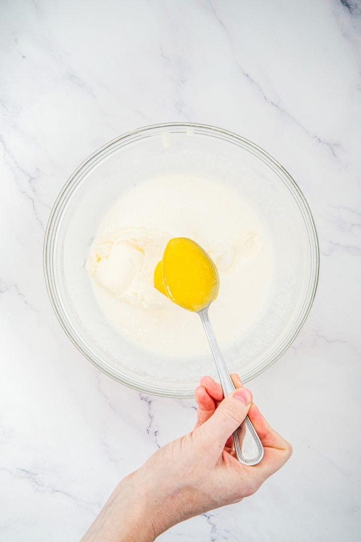 Hand holding spoon of yellow liquid over glass bowl with white ingredients on marble surface, preparing layers for Lemon Cheesecake Trifle Cups.