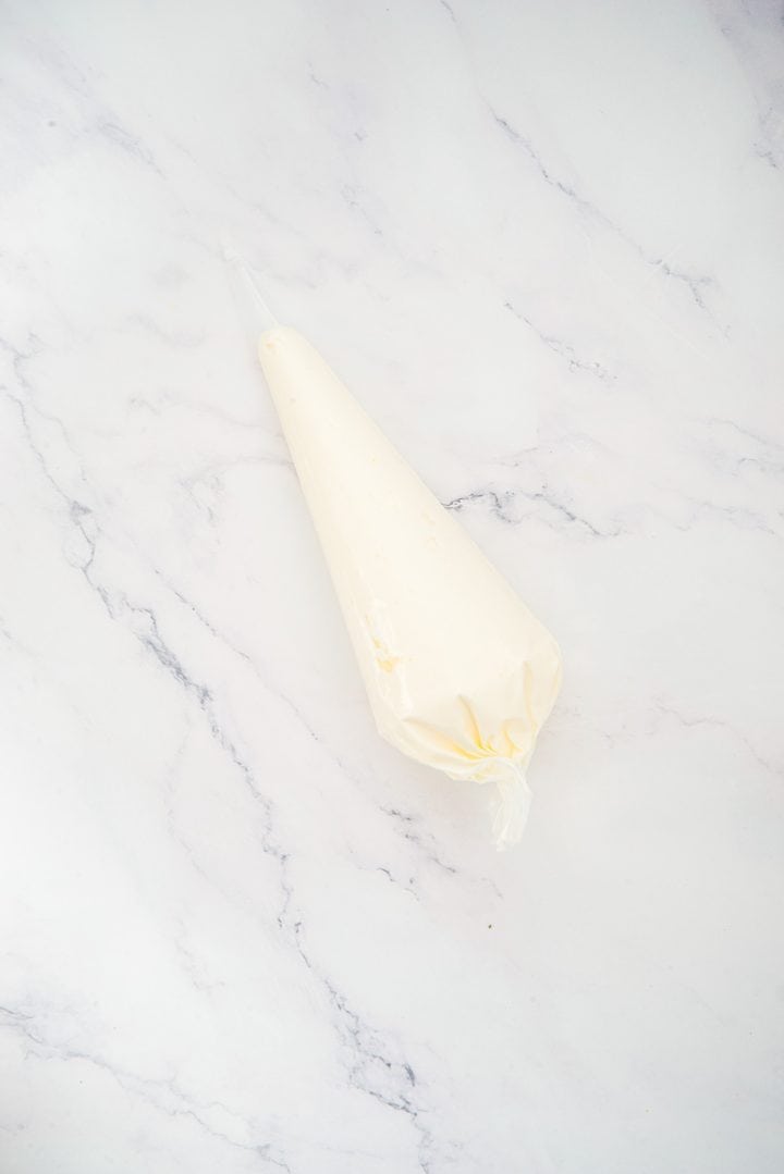 A piping bag filled with white frosting rests on a white marble surface, ready to add the finishing touch to homemade Lemon Cheesecake Trifle Cups.