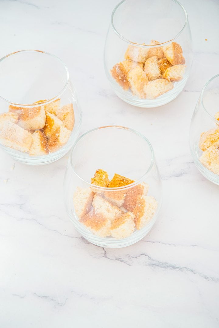 Four glass Lemon Cheesecake Trifle Cups with cubes of bread or cake inside, arranged on a white marble surface.