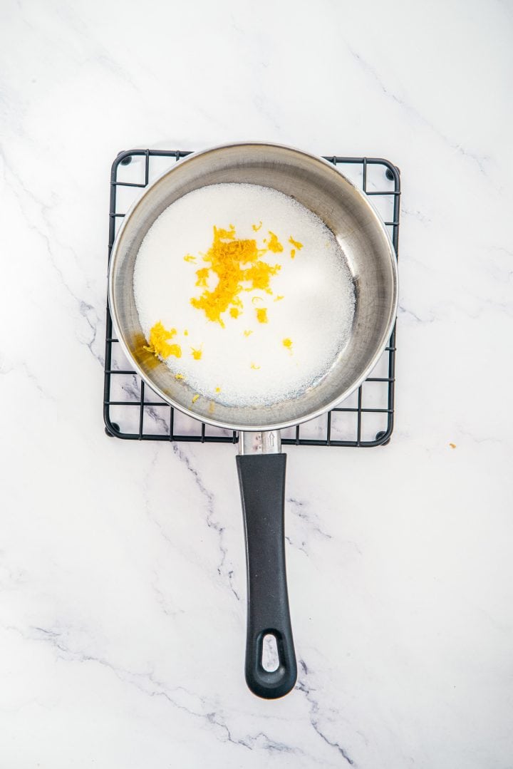 A saucepan with white sugar and lemon zest, perfect for Lemon Cheesecake Trifle Cups, sits on a cooling rack on a white marble surface.