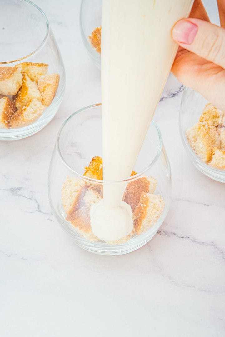 A hand pipes cream into a glass with crumbled cookies on a white surface, creating delicious Lemon Cheesecake Trifle Cups.