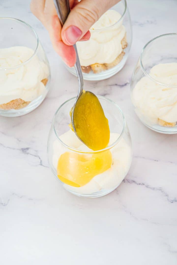 A spoon spreading yellow lemon curd over creamy Lemon Cheesecake Trifle Cups in a glass, with three similar cups nearby.