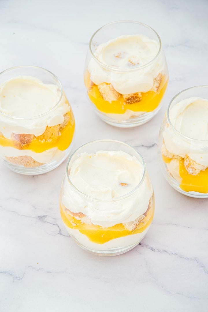 Four glass cups filled with layers of whipped cream, cake pieces, and yellow fruit puree on a white surface, creating delightful Lemon Cheesecake Trifle Cups.