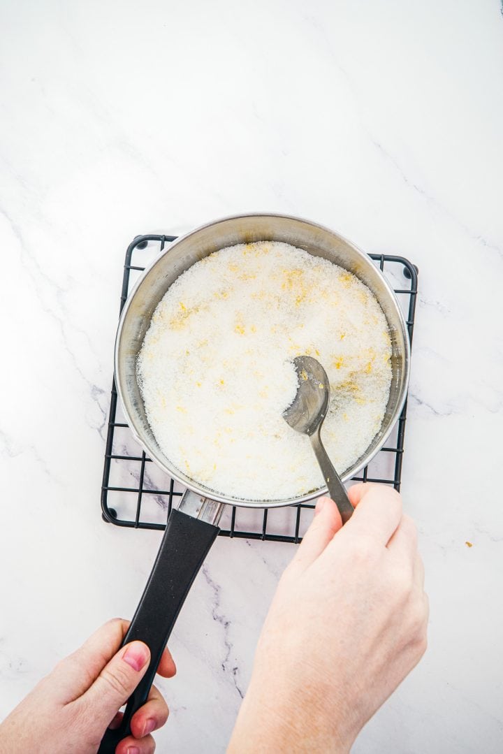 A person stirs a pot of white powder with a spoon on a cooling rack against a white background, preparing the mixture for delicious Lemon Cheesecake Trifle Cups.