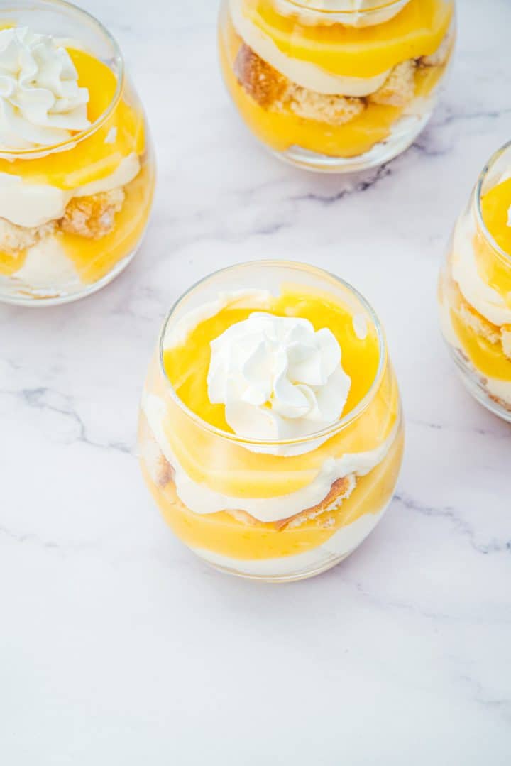 Four Lemon Cheesecake Trifle Cups filled with layered lemon dessert, topped with whipped cream, sit elegantly on a white marble surface.