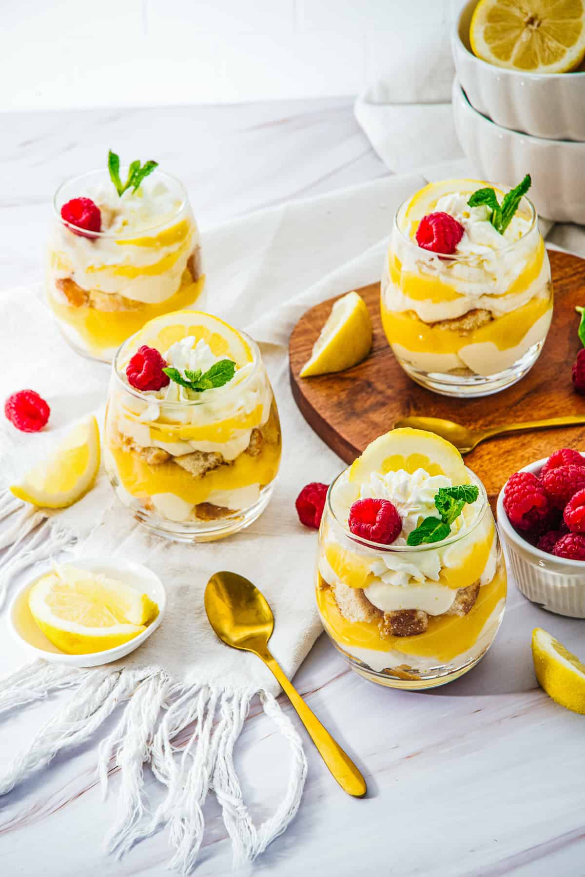 Four Lemon Cheesecake Trifle Cups with whipped cream, raspberries, and mint on a table with fresh lemons and a gold spoon.